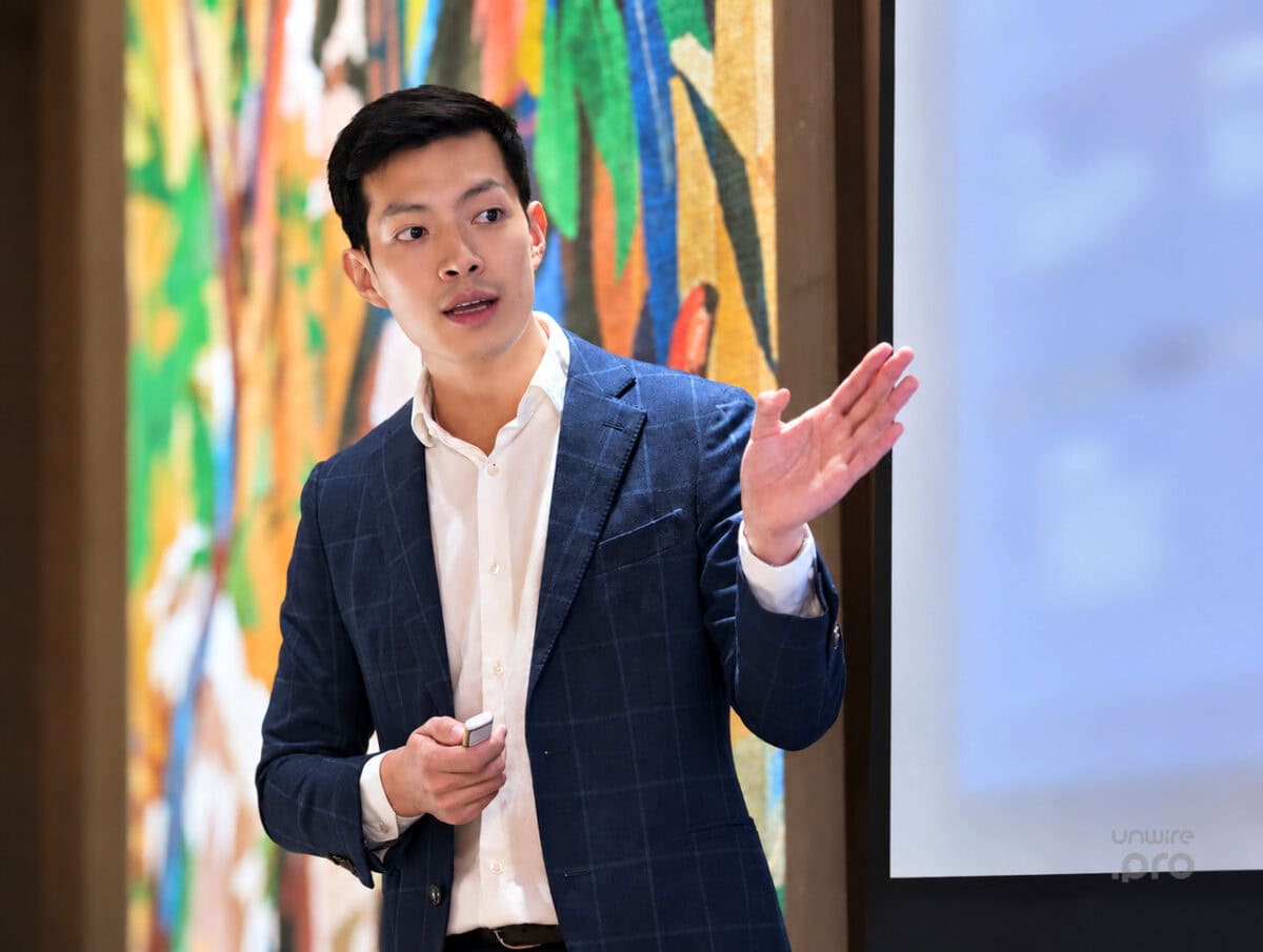 企業 AI 專題講座演講者現場 專注演講的男士穿著深色格紋西裝,手持遙控器在講台前示意,背景是彩色幾何裝飾與投影螢幕.